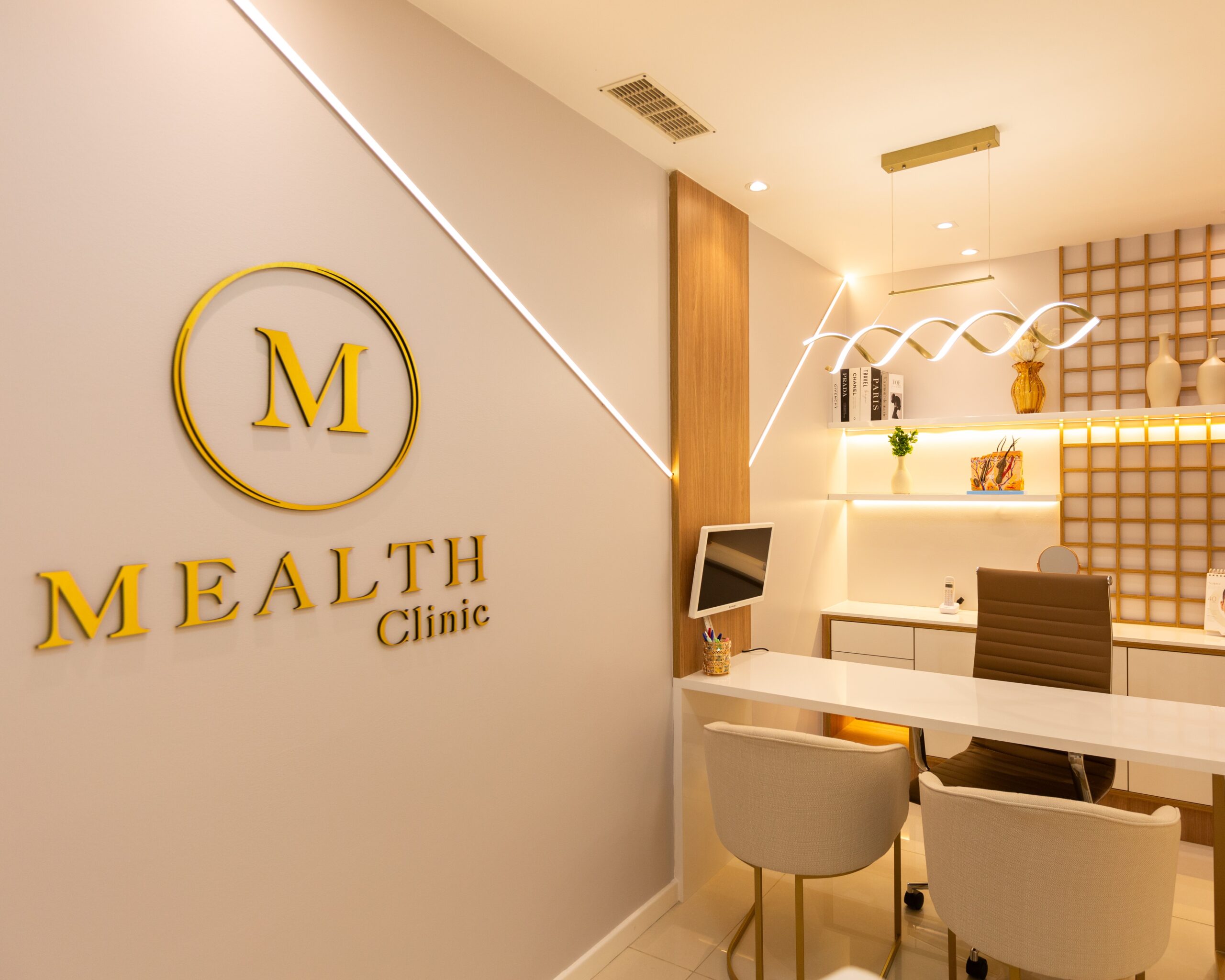 Consultório da Mealth Clinic, clínica dermatológica com foco em estética localizada na Barra da Tijuca e no Cachambi, com o logo, uma iluminação aconchegante e moderna com duas poltronas para pacientes e a cadeira da dermatologista especialista.