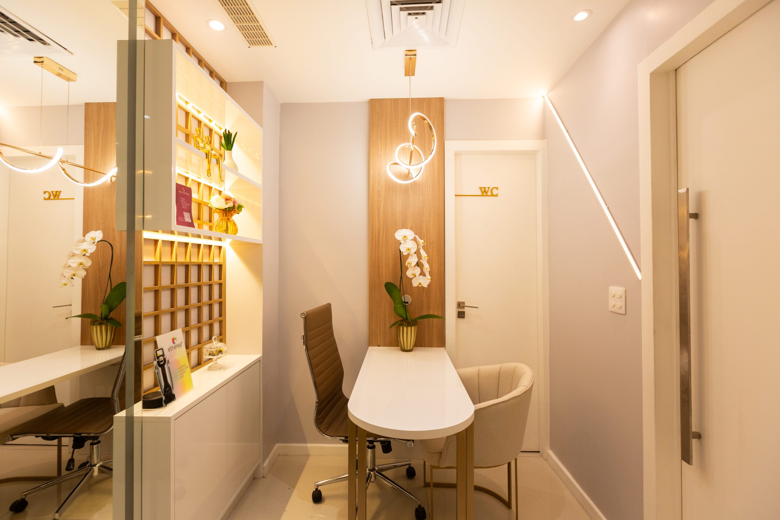 Consultório da Mealth Clinic, de dermatologia estética, localizada na Barra da Tijuca e na Zona Norte do Rio de Janeiro. O consultório tem uma mesa com uma orquídea branca em cima, uma poltrona confortável para o paciente, a cadeira para a médica dermatologista especialista e uma a iluminação aconchegante e moderna.