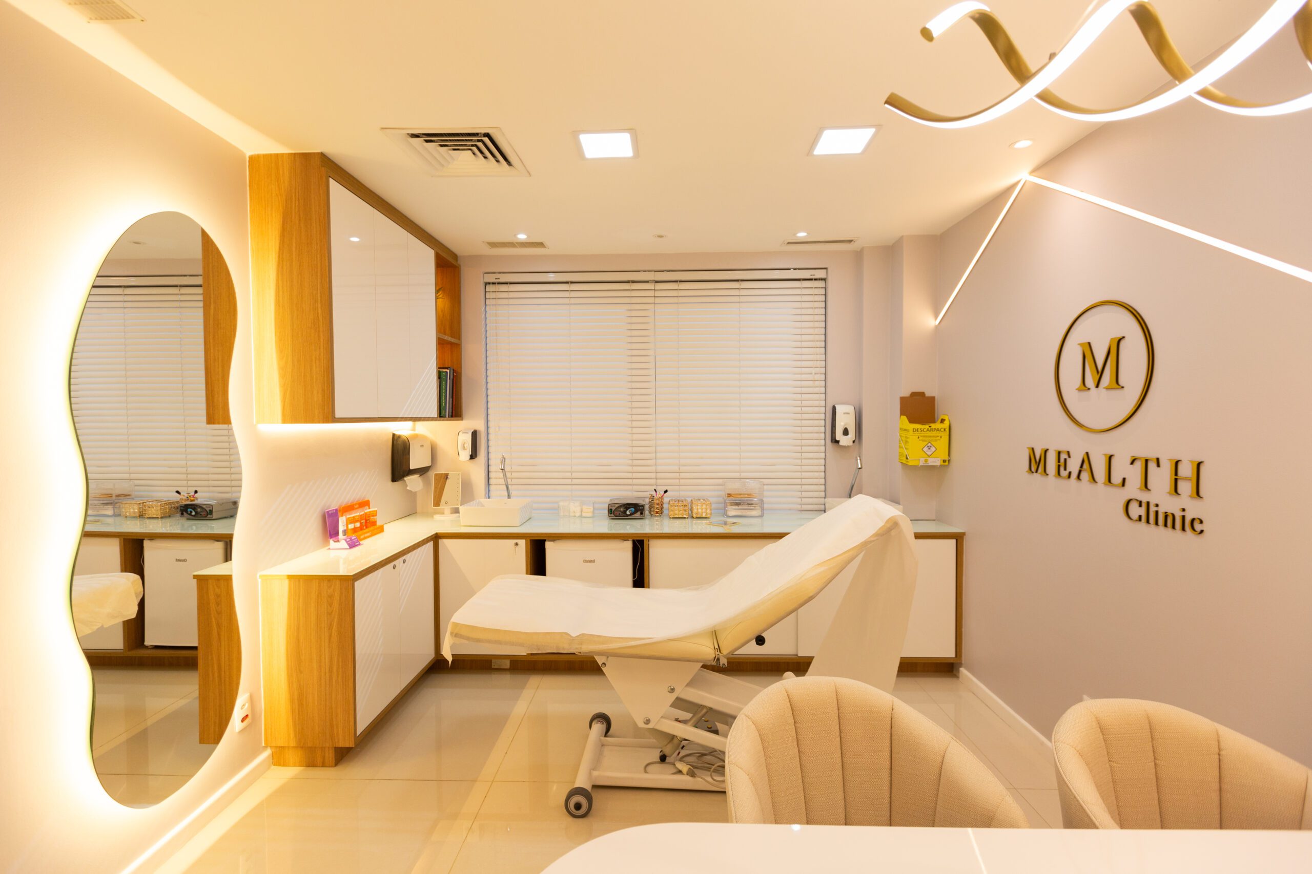 Consultório da Mealth Clinic de dermatologia estética localizada na Zona Norte do Rio de Janeiro e na Barra da Tijuca. O consultório tem duas poltronas confortáveis para os pacientes, um espelho em formato orgânico e com led, uma maca alta e confortável e um armário baixo de vai de uma parede a outra. Além disso, tem seu logo em 3D na parede e uma iluminação moderna e aconchegante.