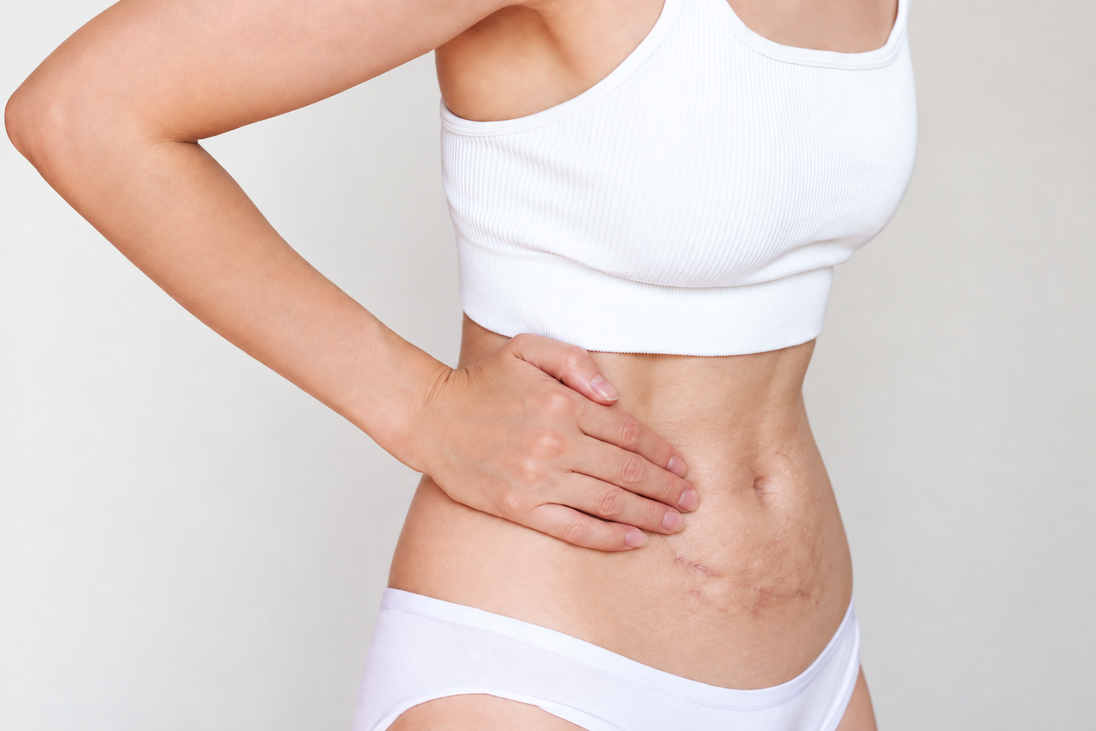 Mulher usando top branco e roupa íntima clara, com a mão apoiada na cintura, destacando a textura natural da pele e marcas corporais em uma fotografia minimalista com iluminação suave.